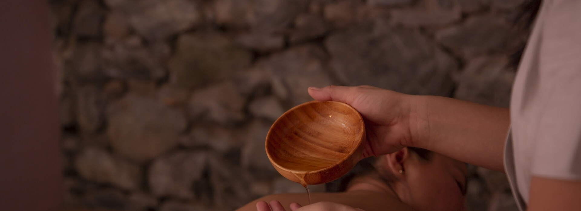 A person in a white shirt pours oil from a wooden bowl onto another person's hand against a blurred stone wall background.