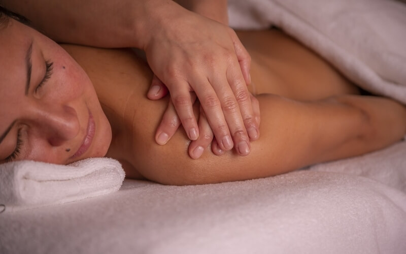 A woman lies on her side receiving a massage, her head on a towel, with a therapist's hand on her arm, exuding relaxation.