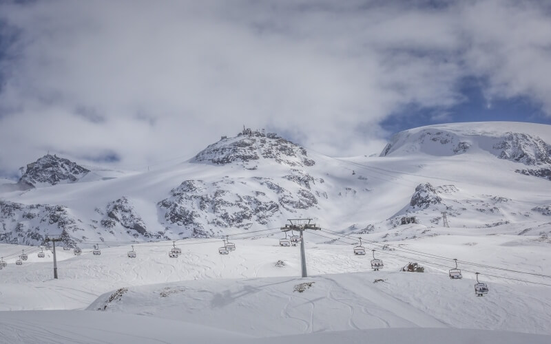 Serene snowy mountain landscape with ski lifts, dark footprints, and majestic peaks under a layer of clouds.