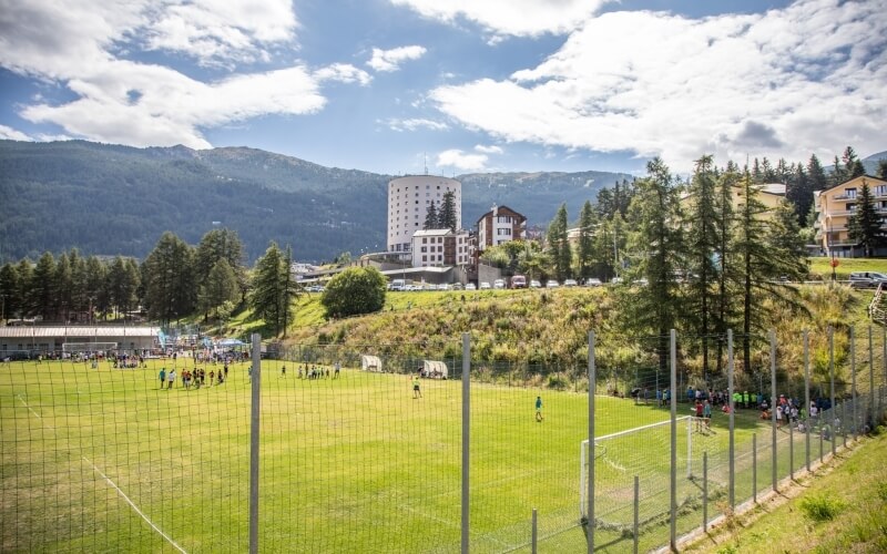 Campo da calcio vibrante circondato da alberi, montagne e un hotel, sotto un cielo blu punteggiato di nuvole bianche.