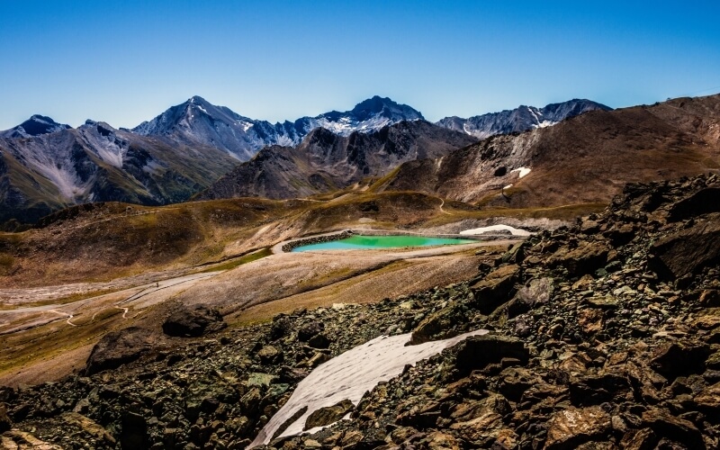 Mountainous landscape featuring a bright blue lake, rocky terrain, and snow-capped peaks in the background.