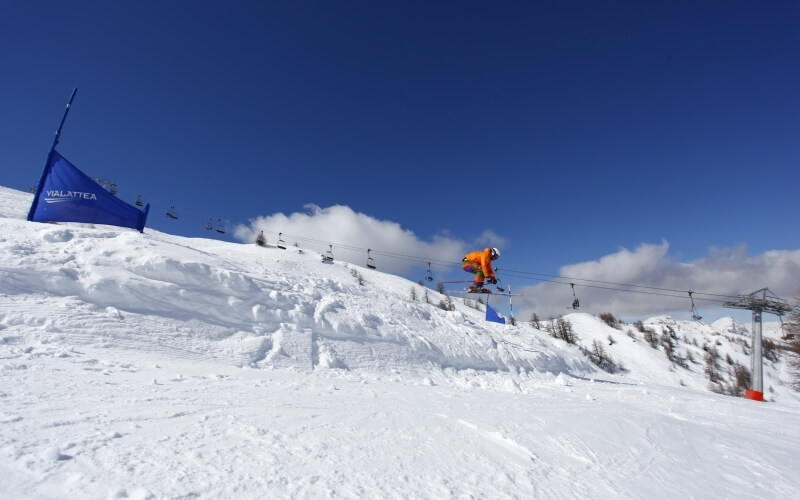 Sciatore in aria su una collina innevata, indossando giacca arancione e casco bianco, con cielo blu sullo sfondo.