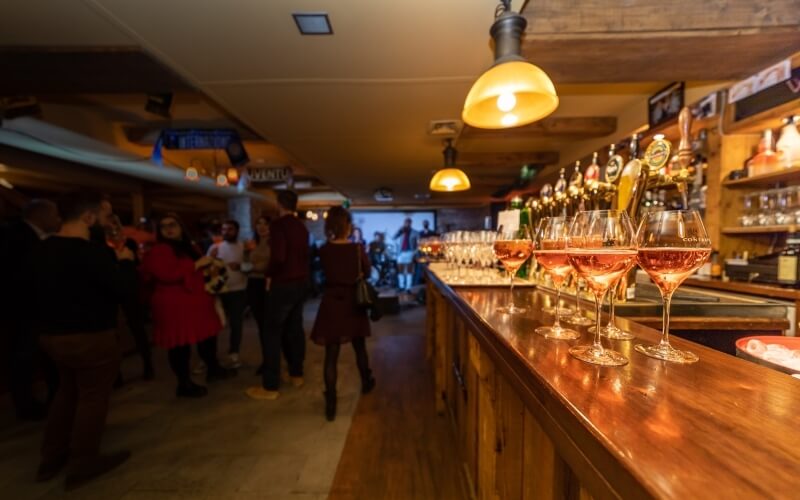 A lively bar scene with a wooden counter, glasses of wine, and warm lighting, featuring people chatting in the background.