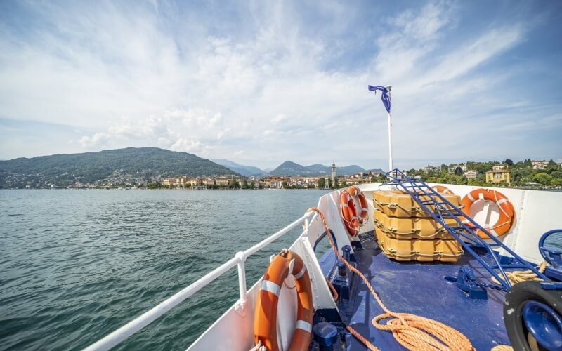 A serene boat on calm water, with a picturesque town and mountains in the background under a blue, cloudy sky.