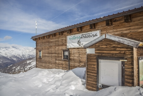 Serene winter scene with a rustic wooden building, "ristorante treccira" sign, snow-covered ground, and snow-capped mountains.