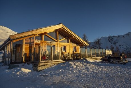 Wooden lodge in snowy mountains with large windows, snowmobile nearby, and tracks indicating recent activity under a clear sky.