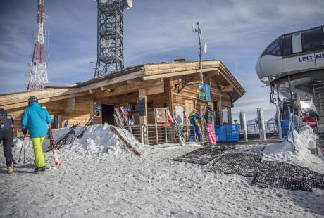 Ski resort scene with a wooden building, skiing equipment, and individuals in winter attire on a snowy ground.