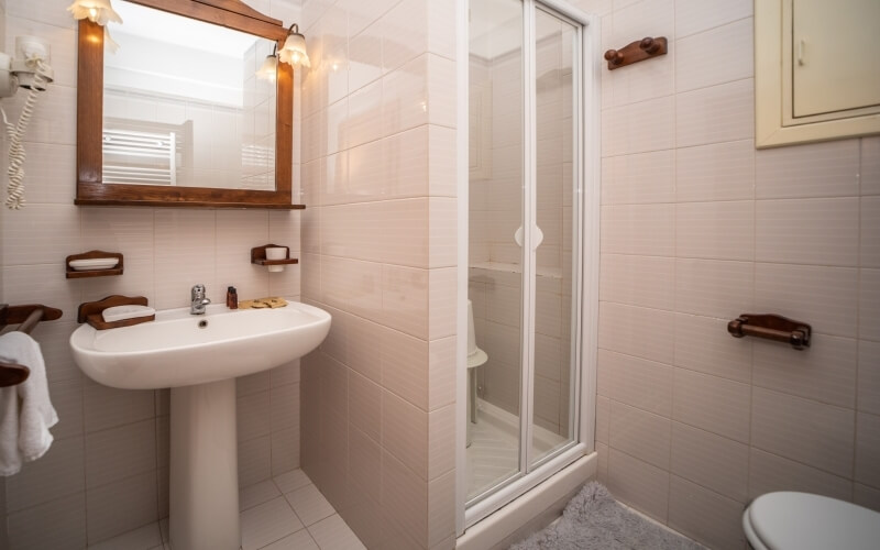 Bathroom with sink, mirror, lights, wooden shelves, toilet, glass shower door, and tiled walls and floor.