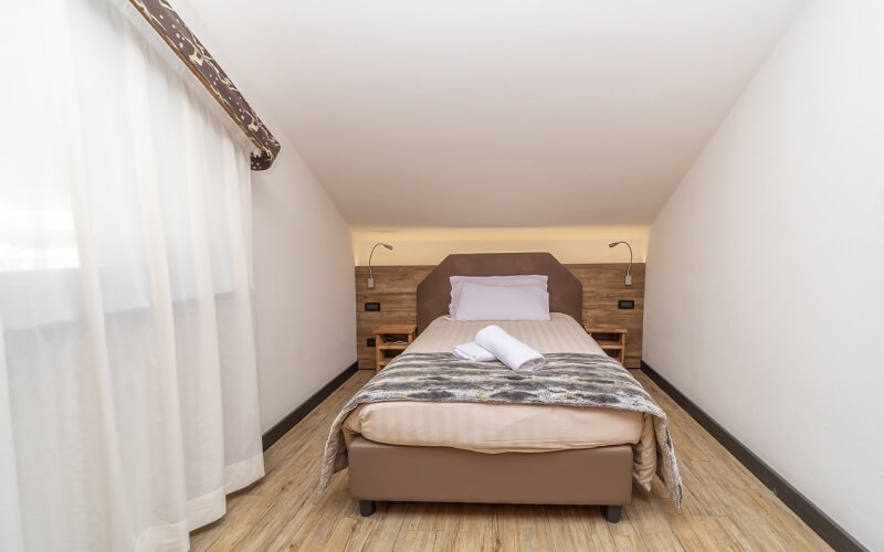 Cozy hotel room with a single bed, nightstands, white curtains, and a warm atmosphere, featuring a patterned blanket and towels.