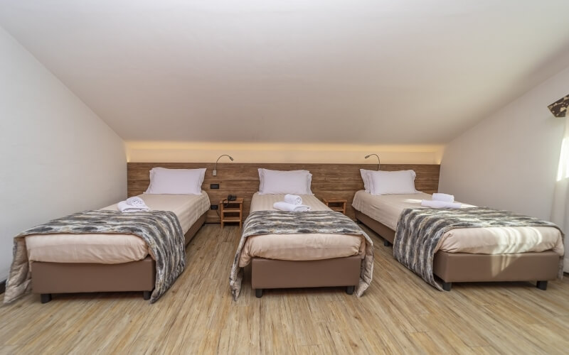 A clean room with three beds, white linens, gray patterned blankets, wooden headboard, and light wood flooring.