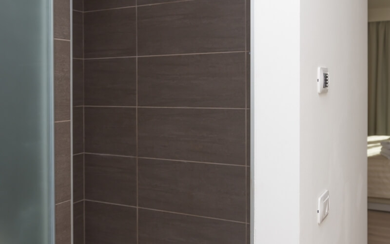 Immagine di un bagno moderno con doccia in piastrelle marroni, parete bianca con termostato e interruttore, e camera sullo sfondo.