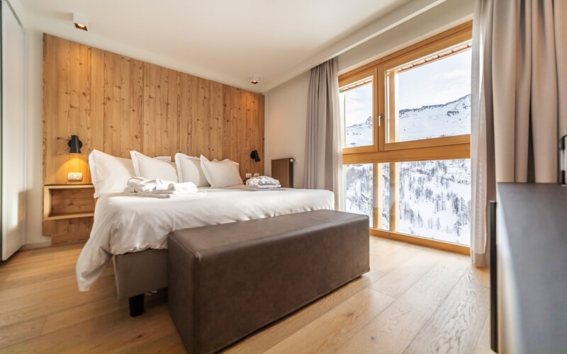 Modern bedroom with a large bed, wooden walls, and a window showcasing snowy mountains, illuminated by natural and lamp light.