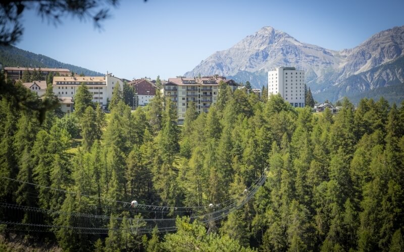 Serene mountainous landscape with a cityscape, featuring buildings among trees and majestic mountains under a clear blue sky.