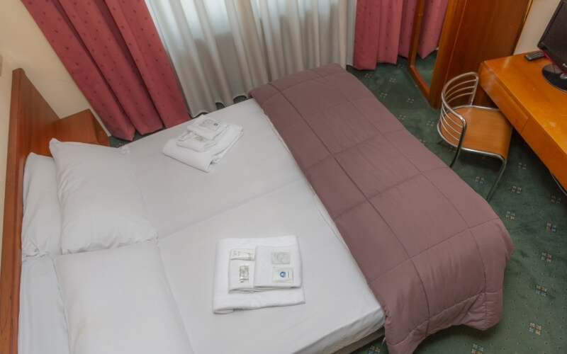 Hotel room with a bed, desk, chair, beige walls, green carpet, and natural light from windows with pink and white curtains.