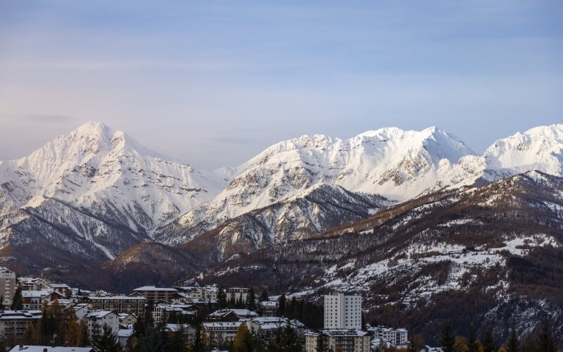 Paesaggio invernale sereno con un piccolo paese ai piedi di montagne innevate e alberi nel primo piano.