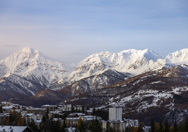 Serene snowy town in the mountains with low-rise buildings, evergreen trees, and snow-covered peaks under a blue sky.