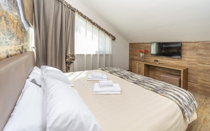 Serene bedroom with a queen bed, light curtains, wall-mounted TV, floral decor, and warm wood flooring for relaxation.