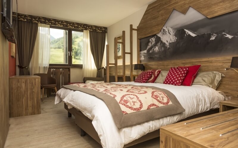 Serene bedroom with a spacious bed, mountain photograph, wooden nightstand, dresser, and brown chairs by a window.