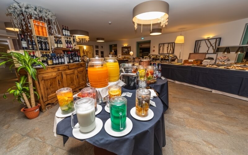 Indoor restaurant buffet with a bar, round tables of drinks, and a food spread, featuring white walls and elegant lighting.