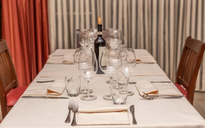 A formal dinner table set with a white tablecloth, six place settings, wine glasses, and a bottle of wine in the background.