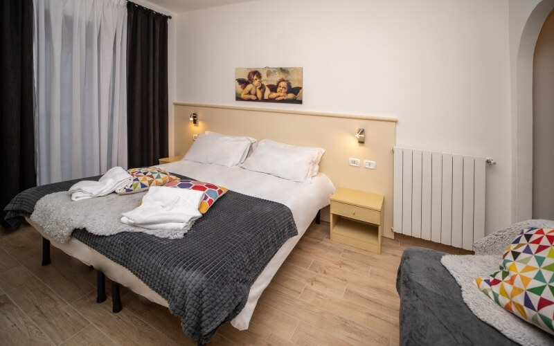 Cozy bedroom with two beds, a nightstand, and a radiator, featuring light wood flooring and dark brown curtains.