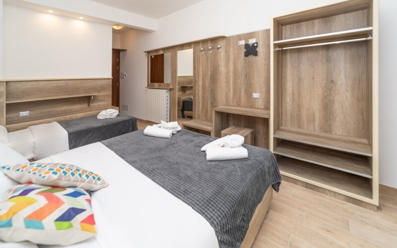 Hotel room with a bed, closet, dresser, and light wood floors; features white and gray comforters and colorful pillows.