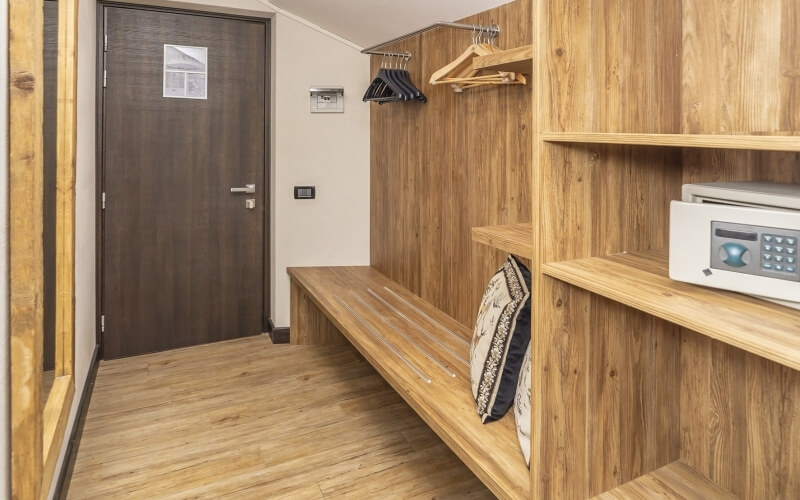 A well-lit room with a wooden door, bench with pillows, and shelves, featuring a white safe on the bottom shelf.