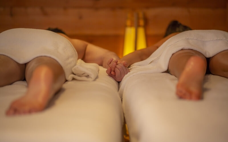 Two individuals relax on massage tables, faces covered with towels, in a serene, softly lit wooden room.