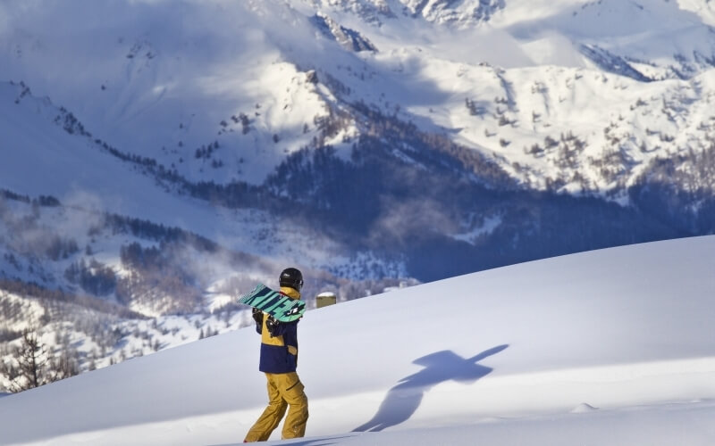 Person in a blue and yellow jacket and yellow pants walks in the snow, carrying a green snowboard with "Ride" on it.