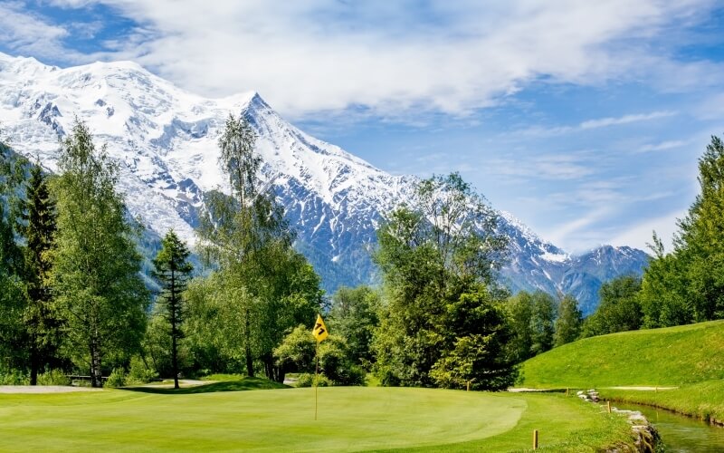 Serene golf course with a green lawn, yellow flag, water body, trees, and snow-capped mountains under a blue sky.