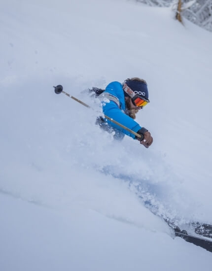 A skier in a blue jacket descends a snowy slope, creating a snow cloud, with trees visible in the background.