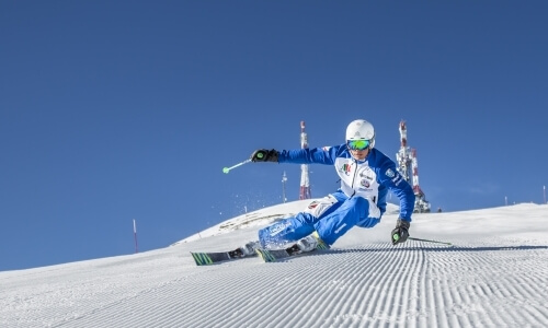 Skier in mid-air on a snowy slope, wearing a blue outfit and white helmet, with ski poles in the background under a blue sky.