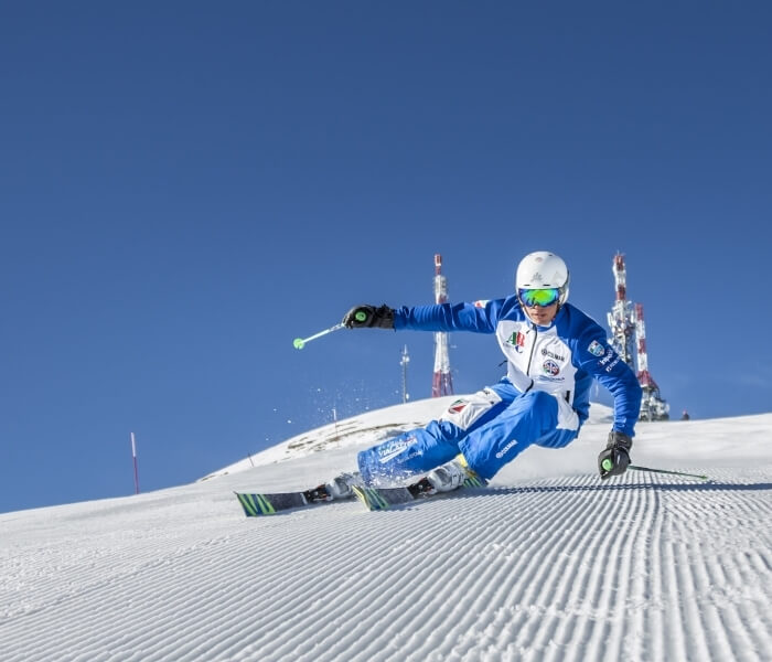 Sciatore in movimento con tuta bianca e blu, occhiali verdi, su fondo innevato con torri rosso-bianco e cielo blu.