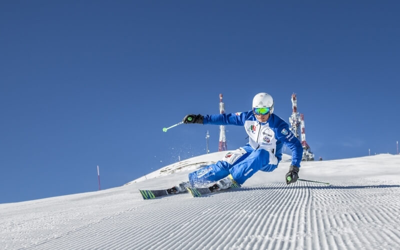 Skier in a white and blue suit glides down a snowy slope, wearing green goggles, with radio towers in the background.