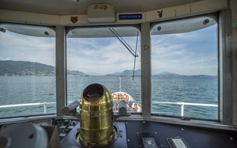 View from a boat's cockpit with controls, a distant boat, calm water, hills, and a serene sky with wispy clouds.