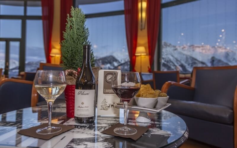 A table with two wine glasses, a bottle, snacks, and a small tree, set against large windows with snow-capped mountains outside.