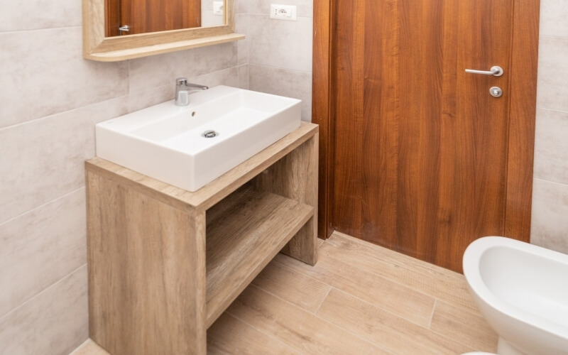 Modern bathroom with white sink, mid-wood cabinet, mirror, wooden door, and beige tile flooring; freestanding bath visible.