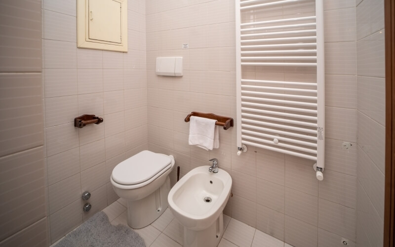 Bagno minimalista con sanitari in porcellana bianca, radiatore e accessori in legno e argento, pavimento in piastrelle bianche.