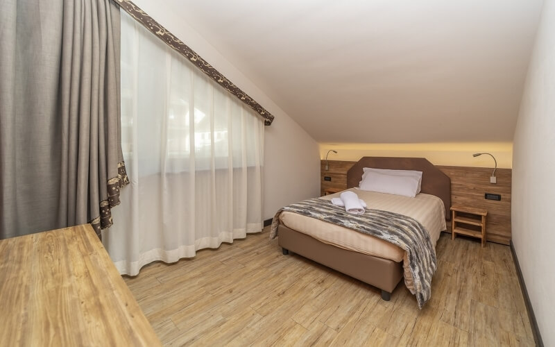 Cozy bedroom with a bed, neutral colors, wooden accents, nightstands with lamps, and sheer curtains letting in sunlight.