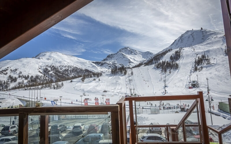 View of a snowy ski resort from a large window, featuring slopes, trees, and a distant parking lot under a partly cloudy sky.