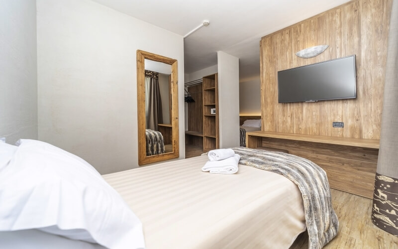 A cozy hotel room featuring a bed with white linens, a mirror, a flat-screen TV, and light wooden flooring.