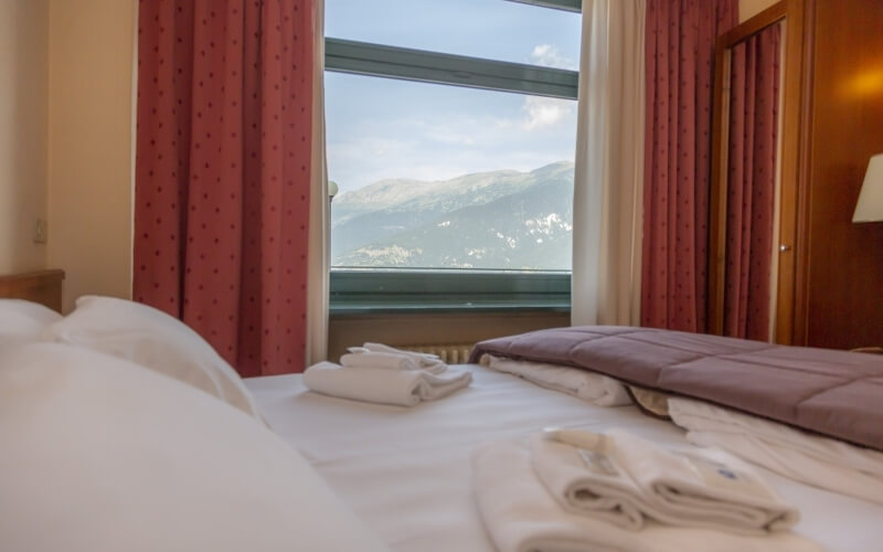 Cozy hotel room with a white bed, red curtains, and mountain view through a large window, featuring folded towels and a purple blanket.