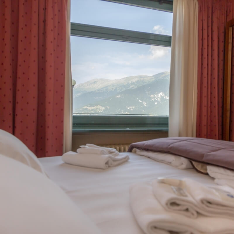 Hotel room with a large window, white bed, red curtains, and a view of snowy mountains outside.