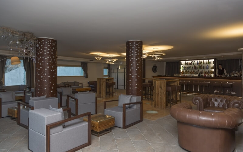 A well-lit lounge with a bar, brown leather couch, gray chairs, and decorative pillars, creating an inviting atmosphere.