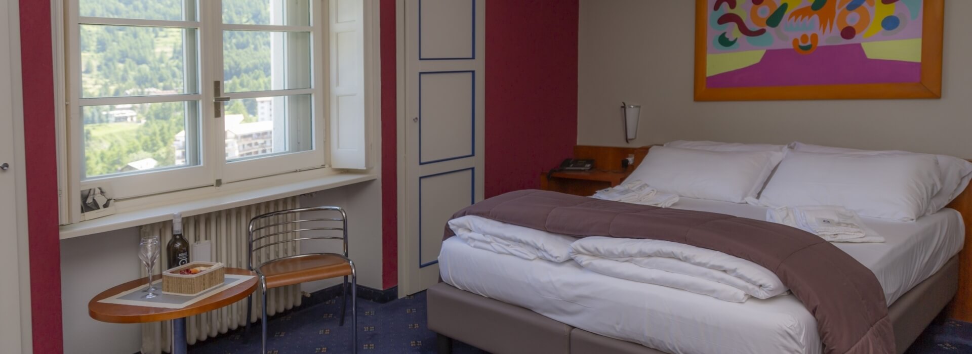 Alpine hotel room with French doors, a double bed, wine tray, and colorful walls, overlooking a town square.