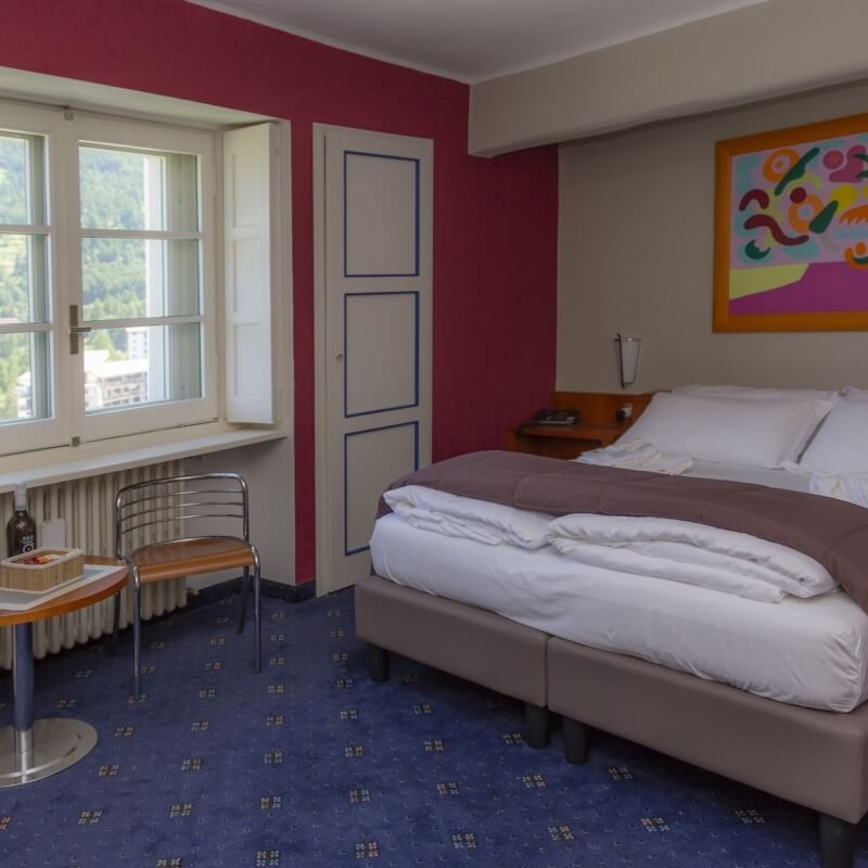 Cozy hotel room with a double bed, window view, desk, and small round table with chairs, featuring red and beige walls.