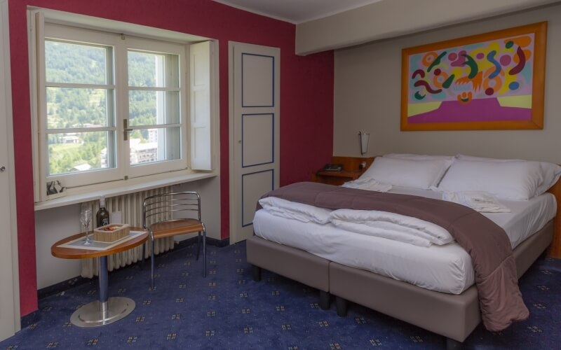 A cozy bedroom with a gray bed, white comforter, brown nightstand, blue patterned carpet, and red-beige walls.