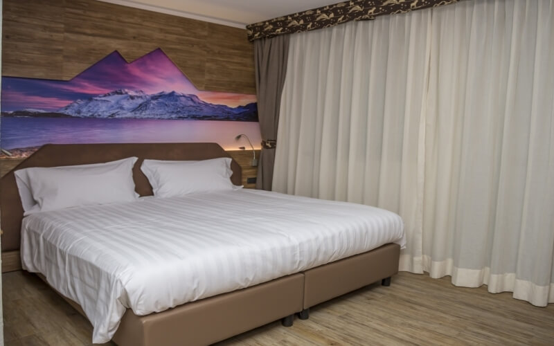 Serene bedroom with a large bed, mountain mural at sunset, sheer curtains, and warm lighting, creating a calm atmosphere.