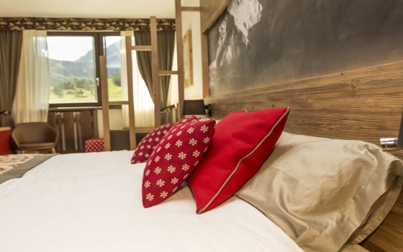 Serene bedroom with a bed, red and white pillows, wooden headboard, window view of mountains, and cozy ambiance.