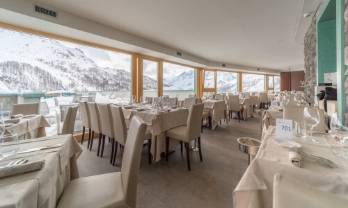 Una sala da pranzo elegante con tavoli ben apparecchiati e vista su montagne innevate attraverso una grande finestra.